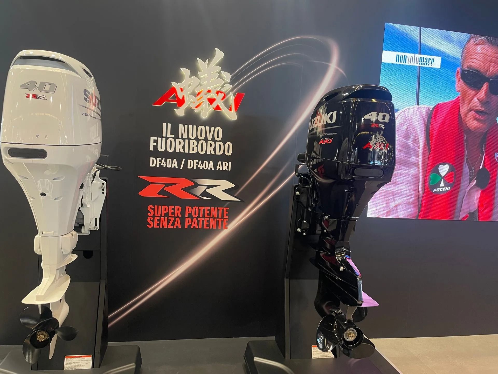 Due motori fuoribordo Suzuki in esposizione: uno bianco e uno nero. Sullo sfondo, testo in italiano e un uomo con un giubbotto di salvataggio su uno schermo.