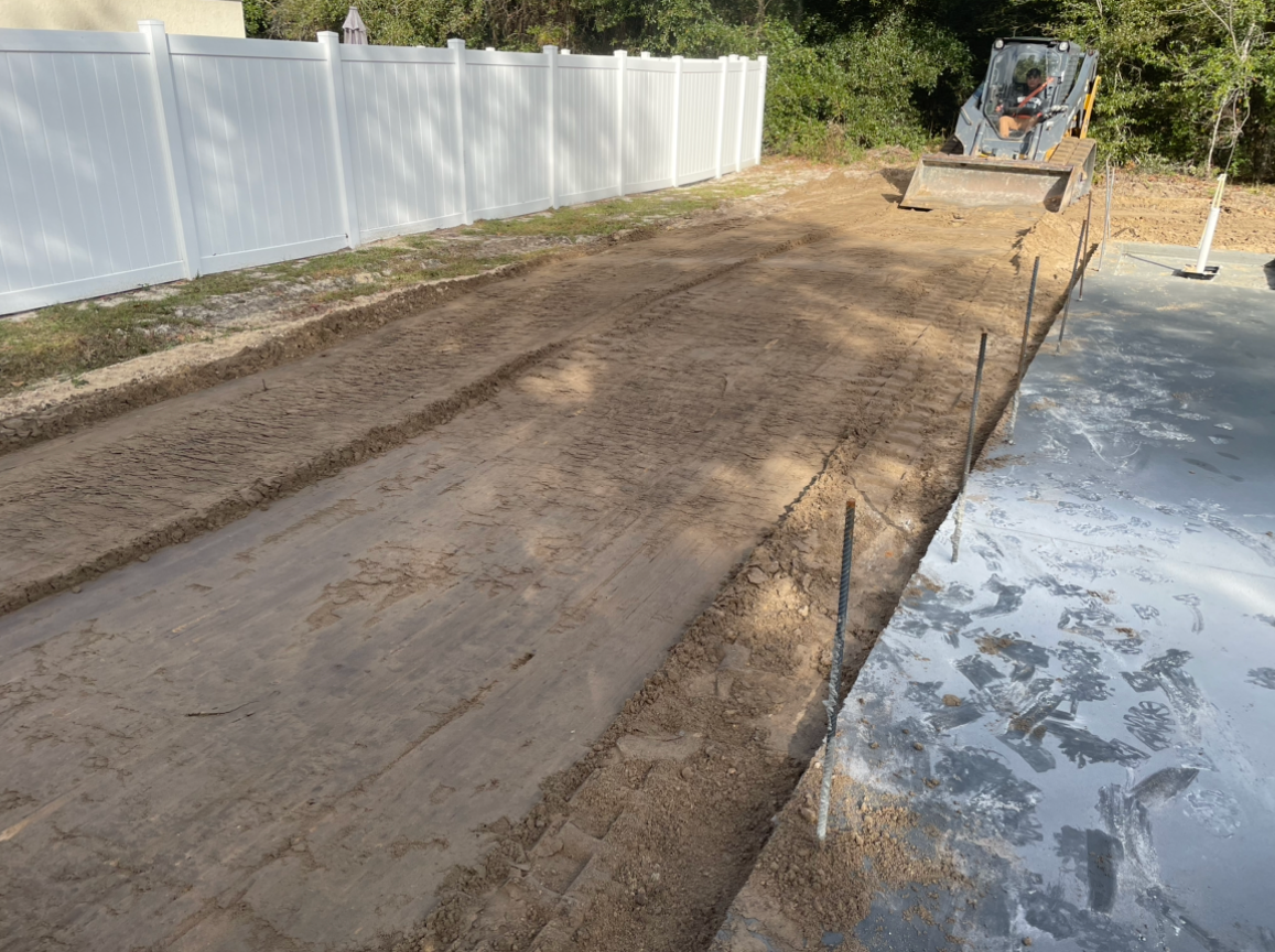 A bulldozer is moving dirt in a driveway next to a white fence. Project done by C1 Foundations