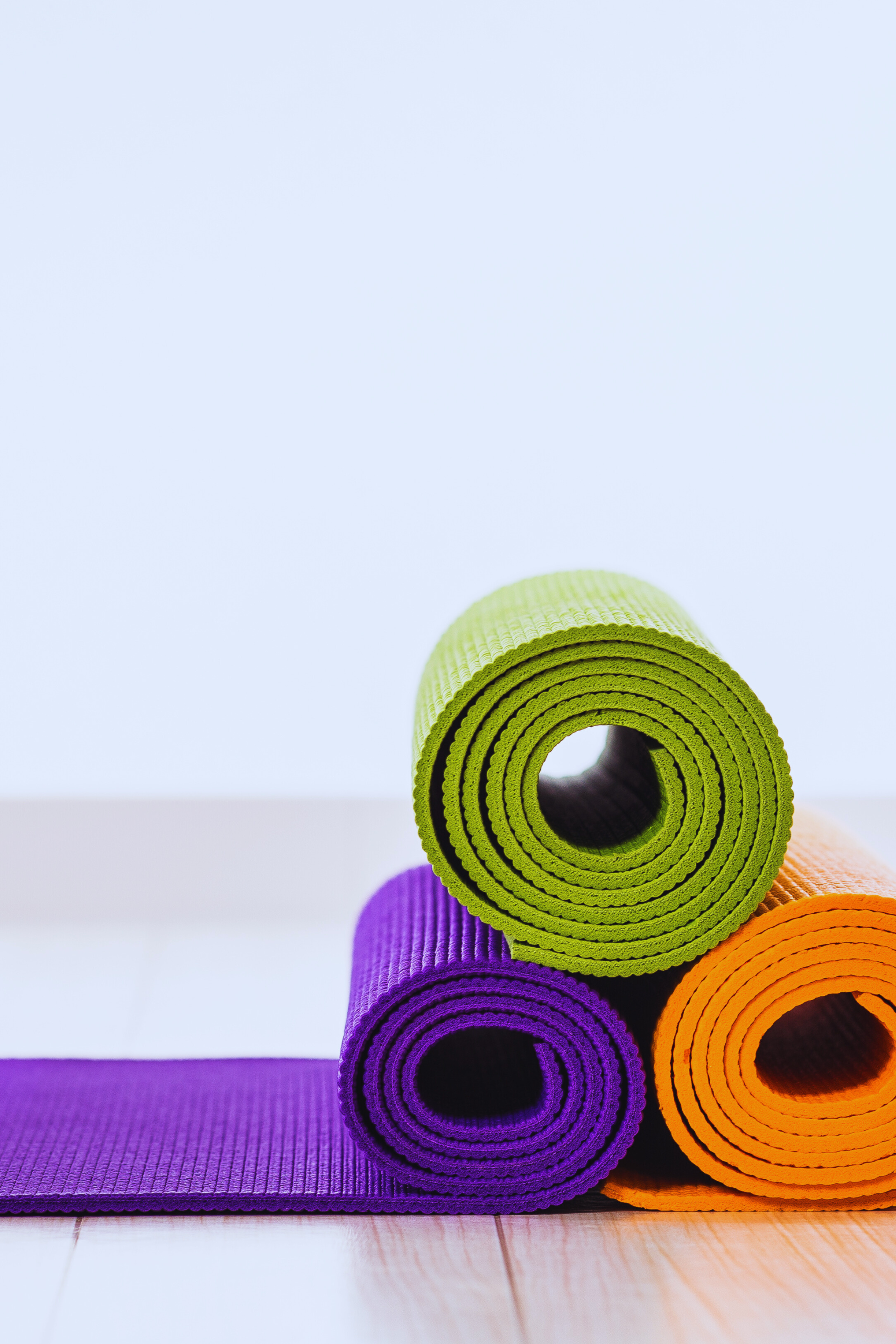 Three rolled-up exercise mats in green, purple, and orange stacked on a light wooden floor against a plain wall.