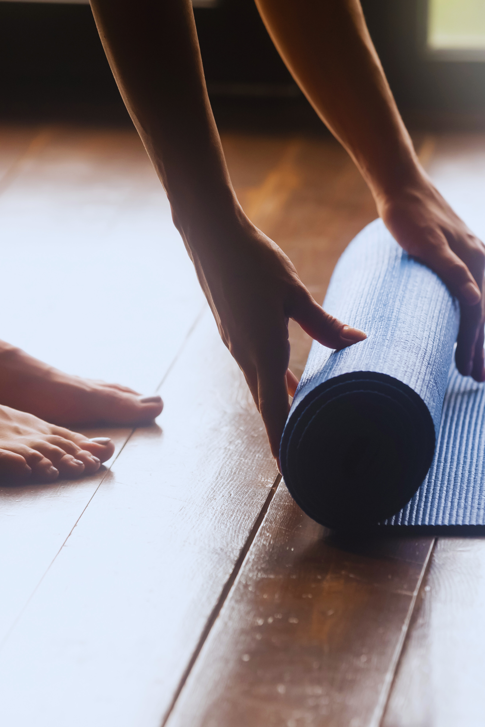 A person's hands rolling up a blue yoga mat on a wooden floor, with bare feet visible nearby.