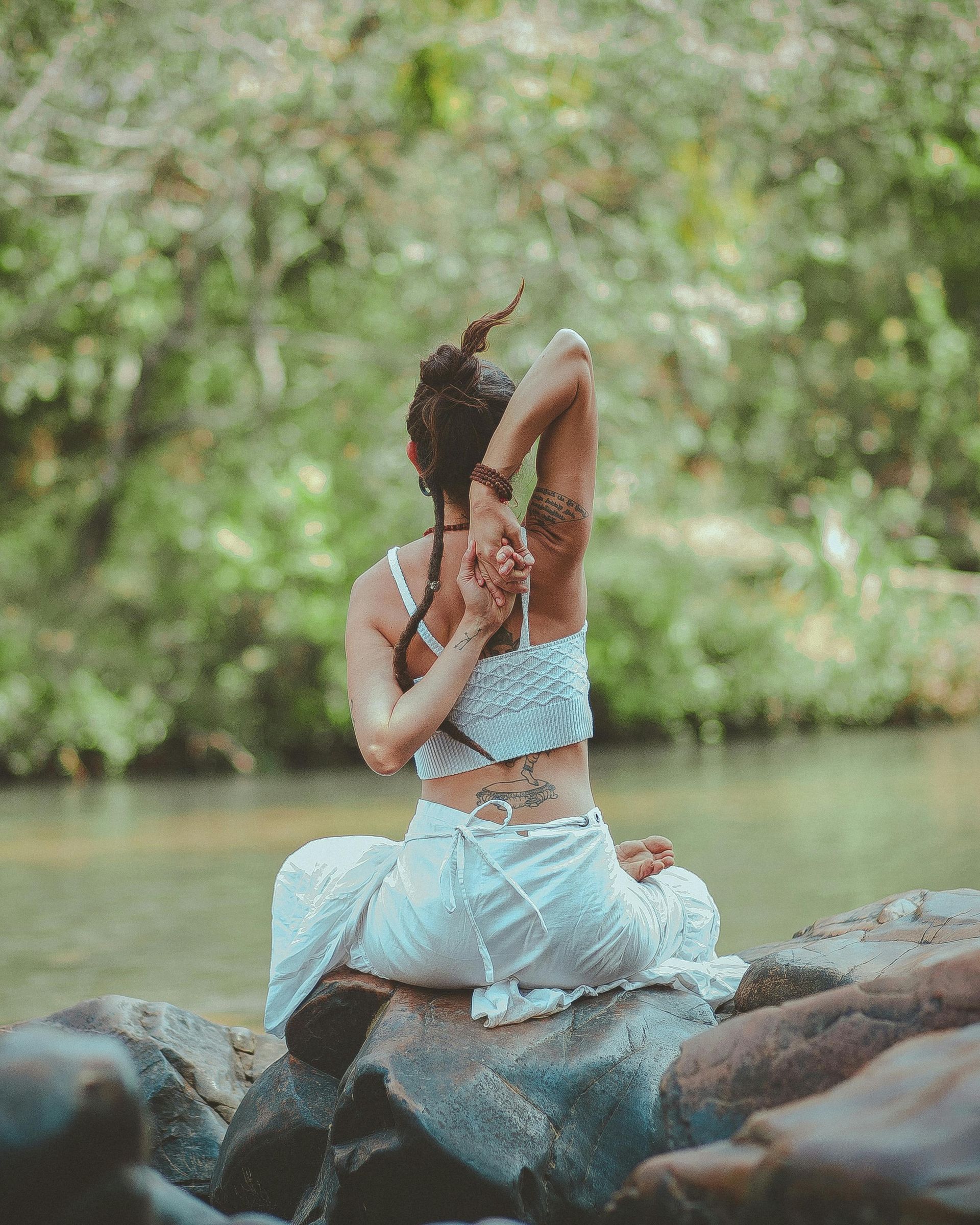 A person with braided hair sits on rocks by a river in a meditative pose, arms clasped behind their back.