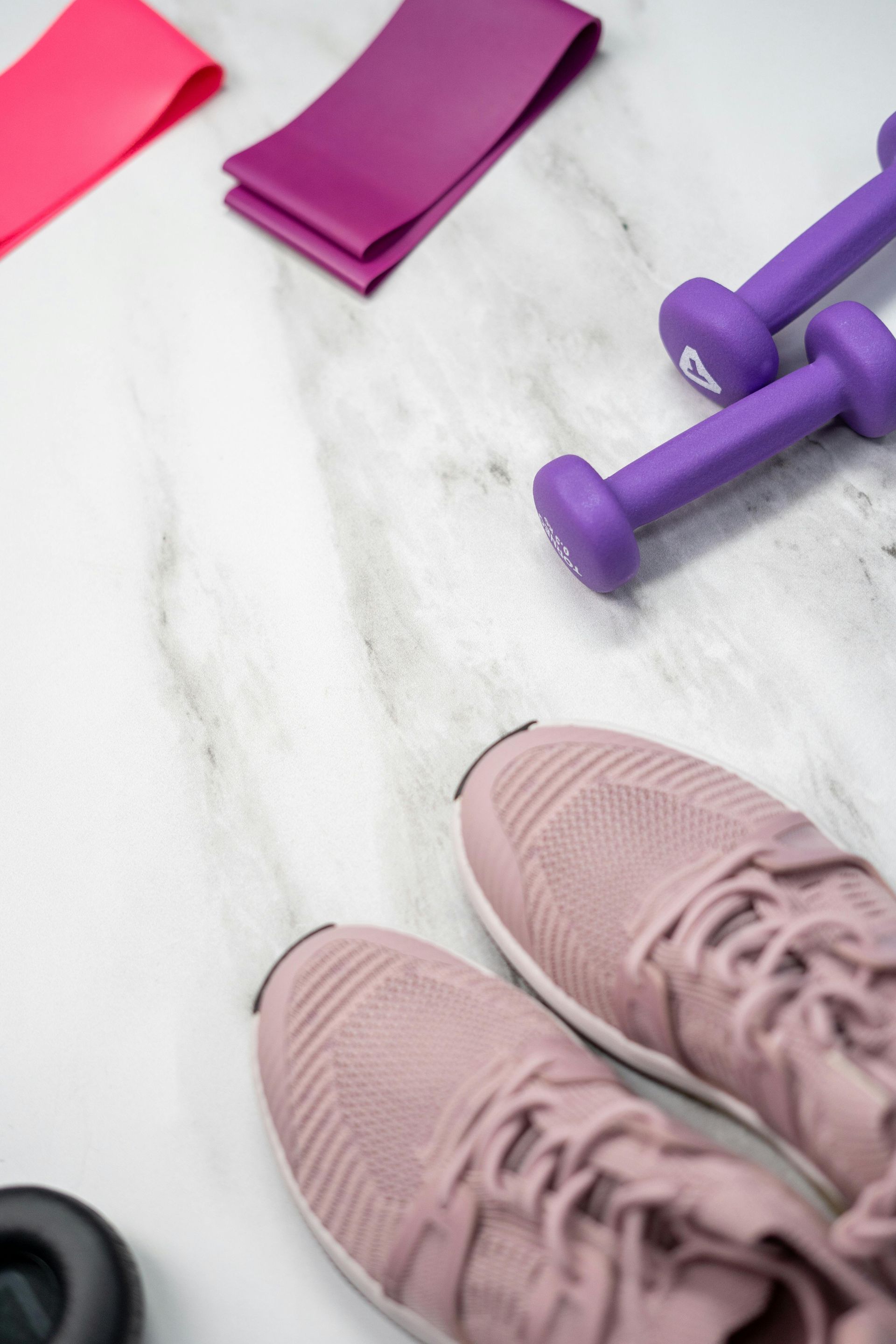 Pink sneakers, purple dumbbells, and resistance bands arranged on a light-colored marble surface.