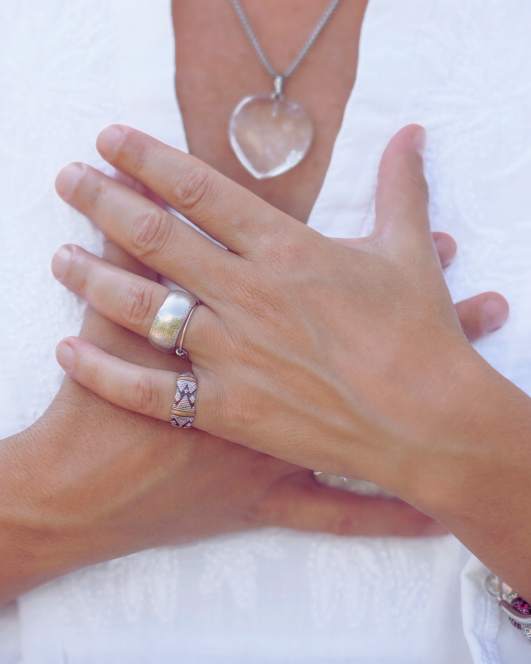 Hands crossed over the chest wearing rings, with a heart-shaped pendant visible on a chain against a white background.