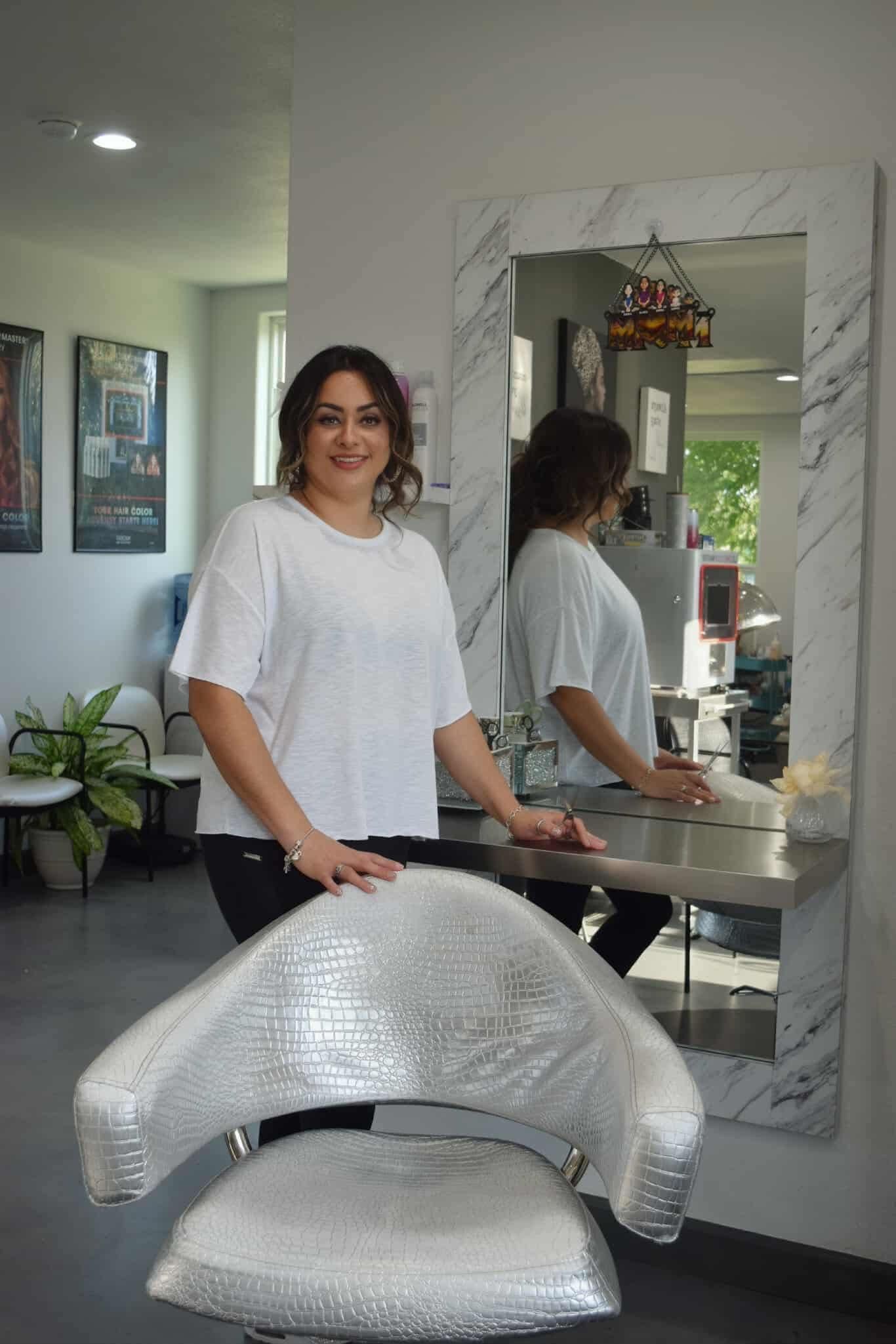 Woman in white shirt stands in salon, hand on silver chair, mirror behind.