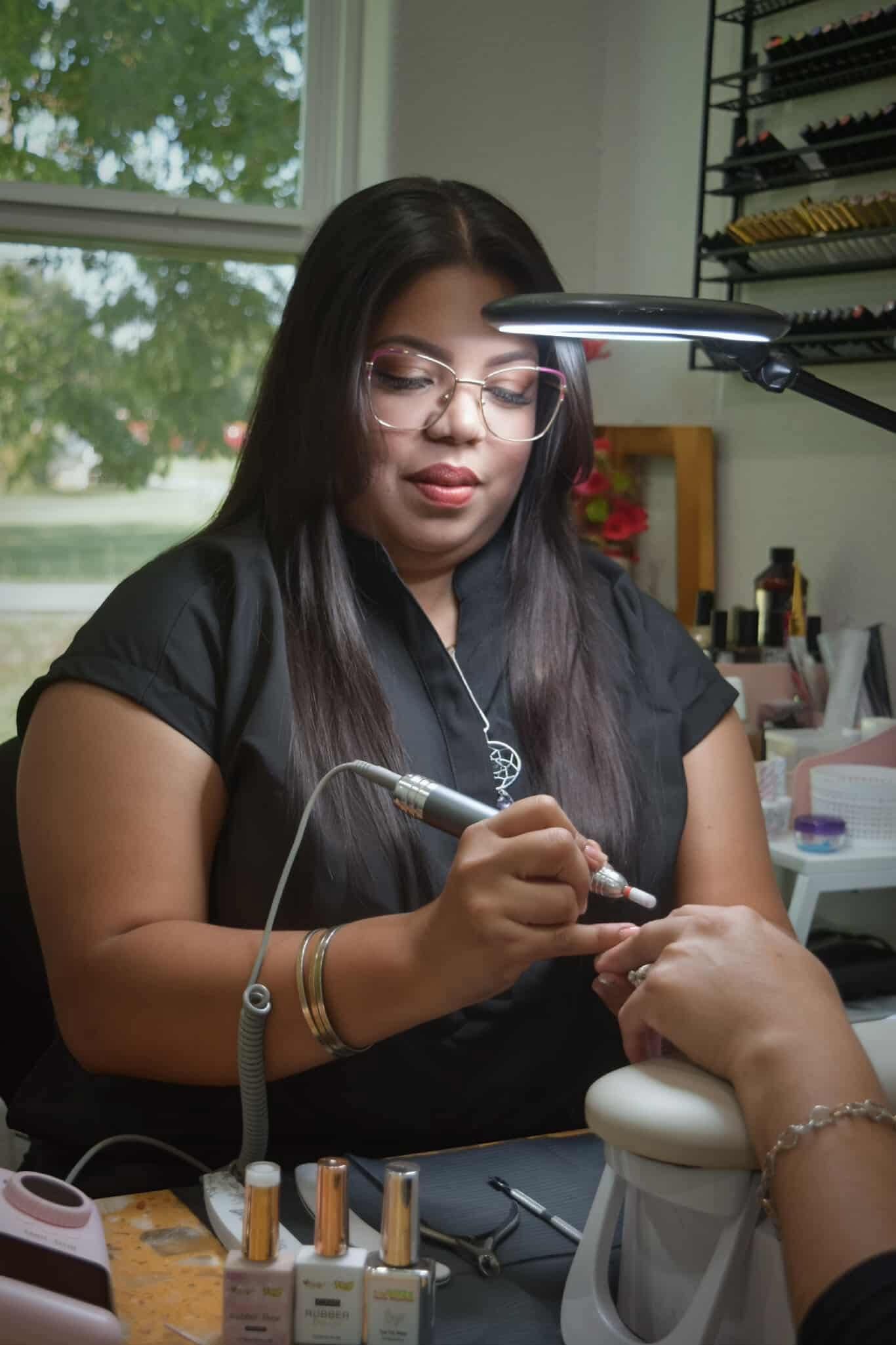 Woman doing a manicure, using a drill. She is wearing glasses, a black shirt, and is in a well-lit salon.