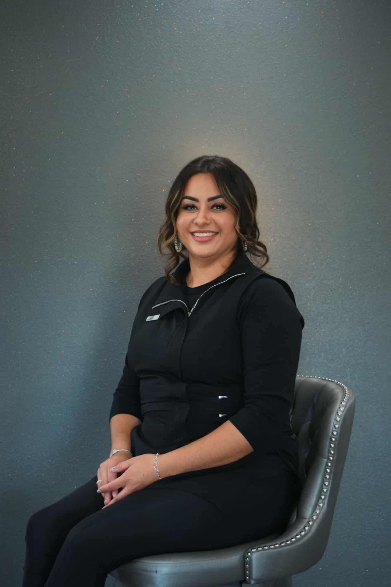 Woman with brown hair in black clothing smiles, sitting on a grey chair, against a grey background.