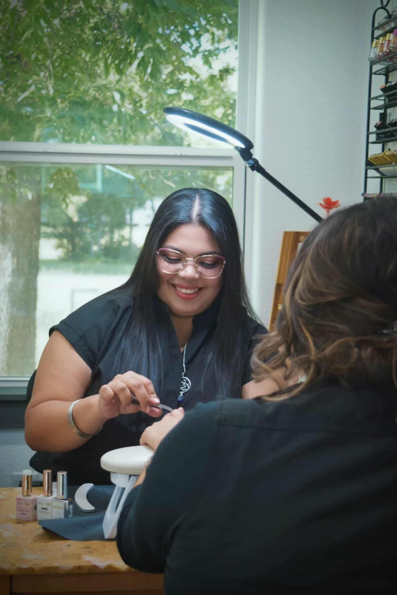 Woman smiles, giving a manicure to a client, next to a window with natural light.