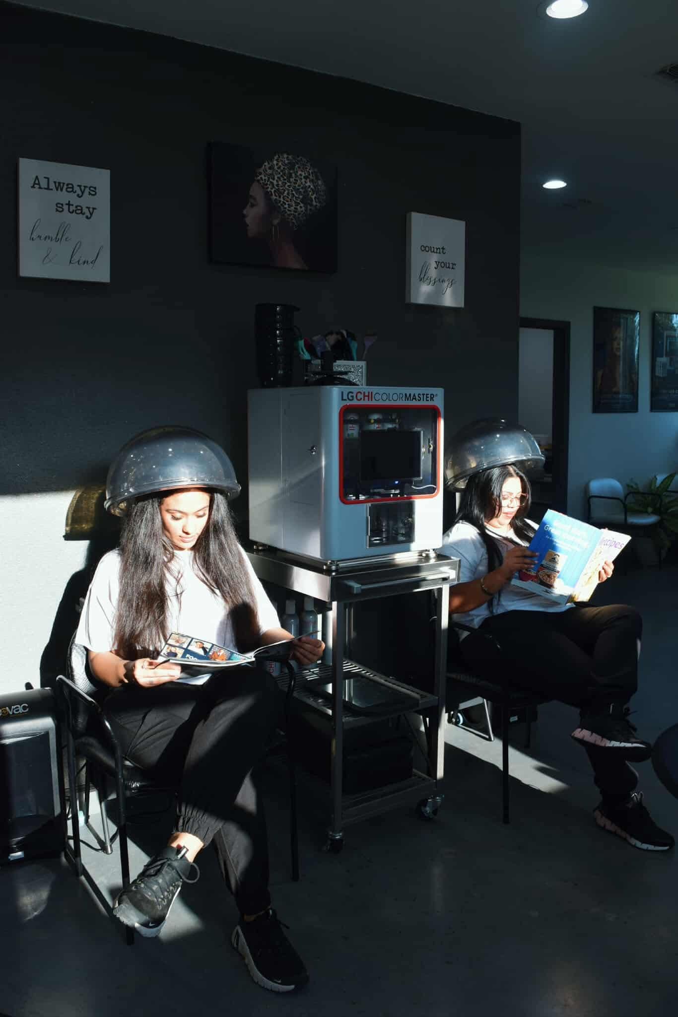 Two people under hair dryers in a salon. Sunlight, dark walls, and posters are visible.