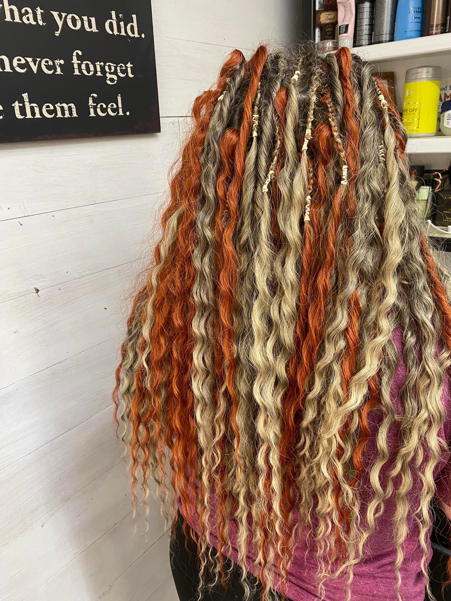 Woman with long, multi-colored, curly braids against a white wall; some bottles on a shelf in background.