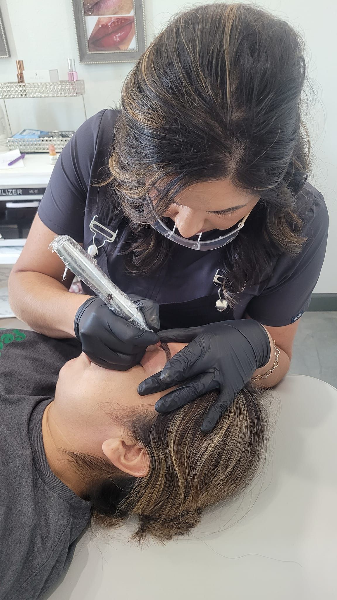 Woman with black gloves using a tool on a man's eyebrow; both are wearing masks, possibly in a beauty salon.