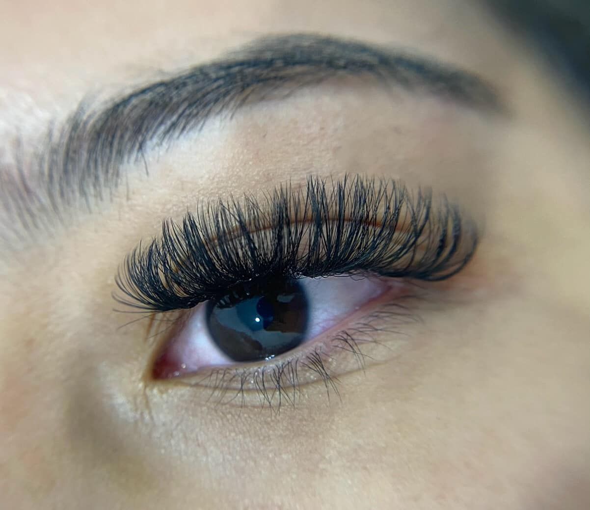 Close-up of an eye with long, black eyelash extensions. The eye is brown and has a neatly groomed eyebrow.