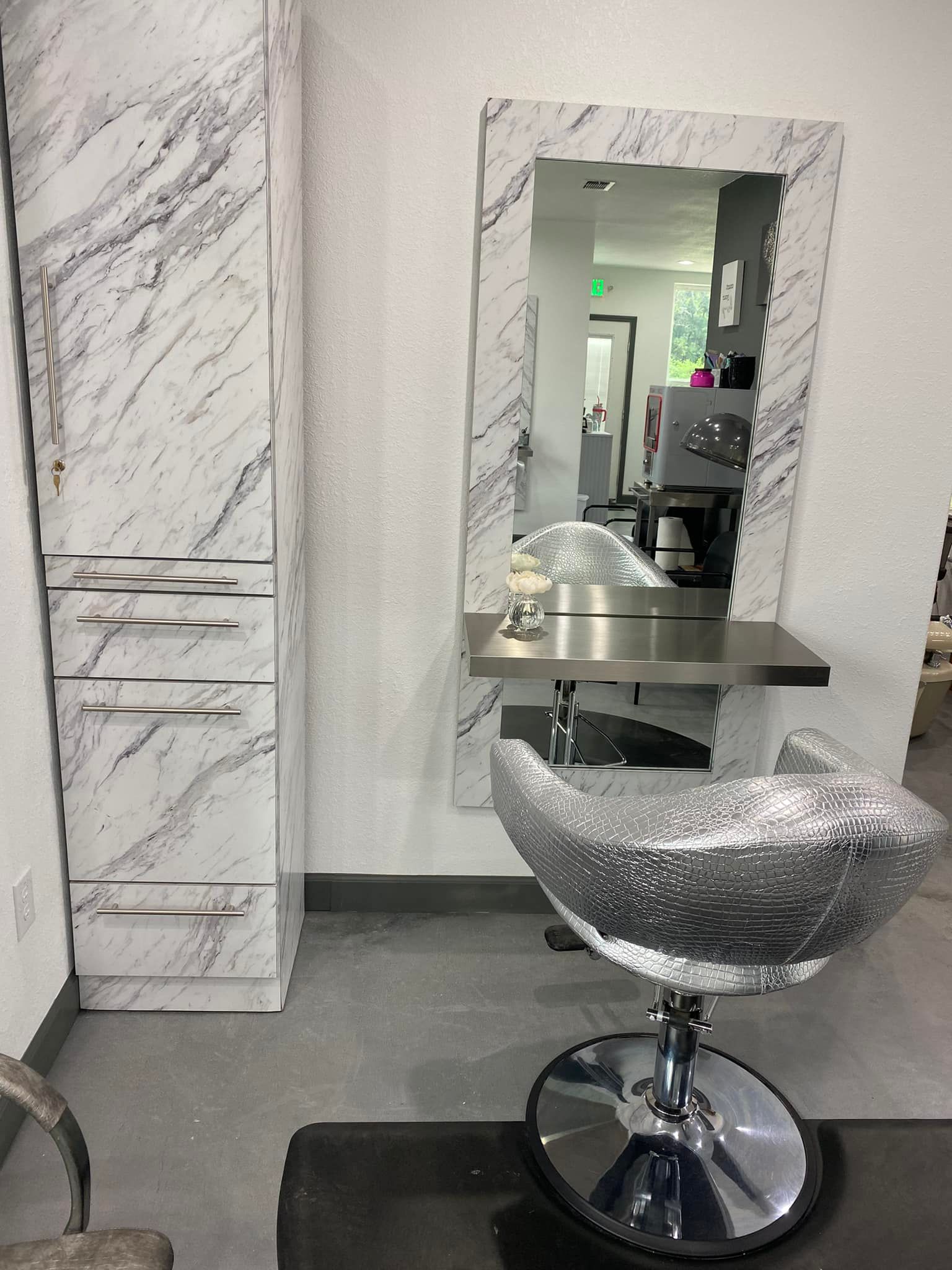 A hair salon station with a marble-look storage cabinet, mirror, stylist chair, and stainless steel table.