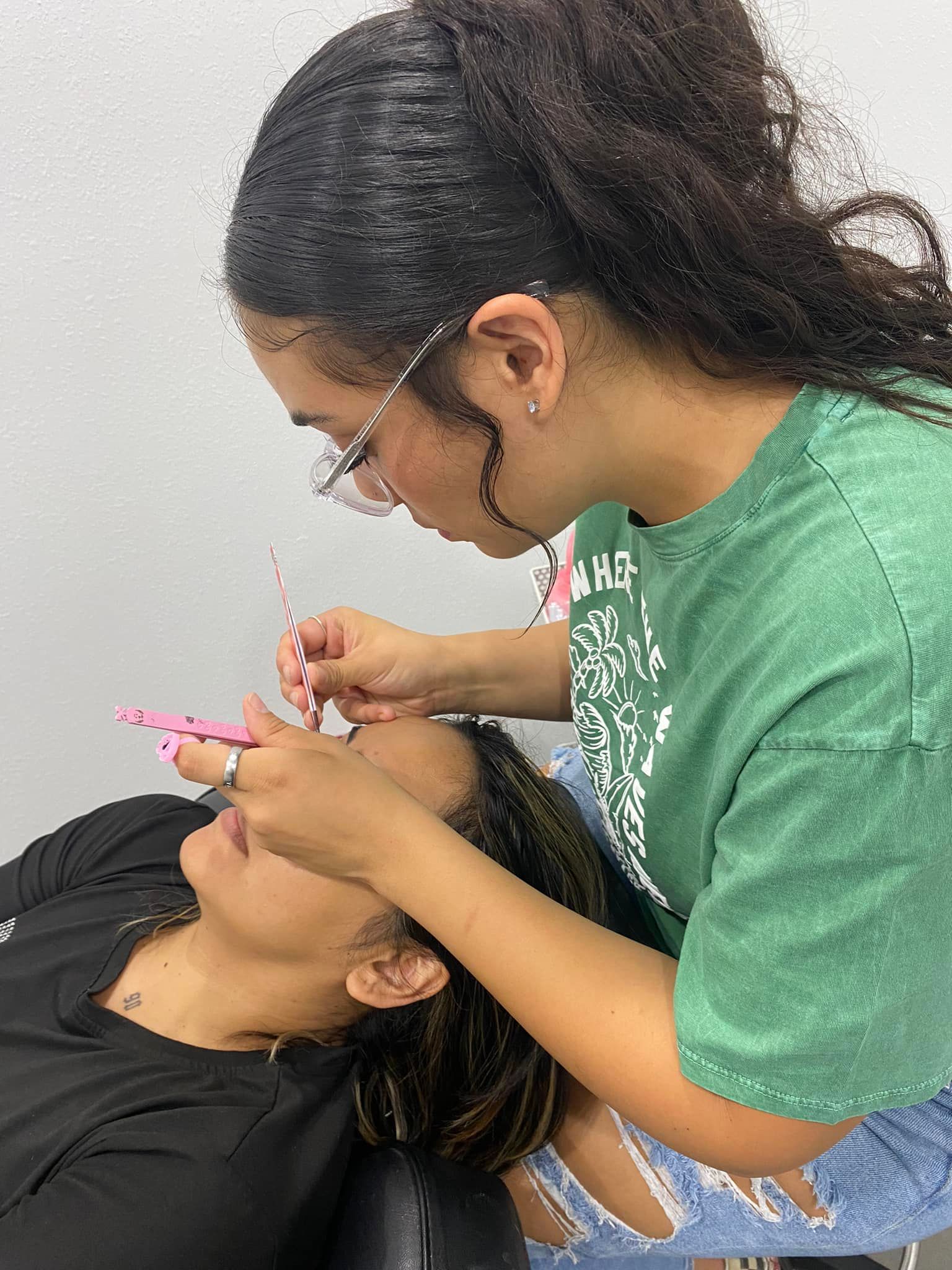 Woman applying eyelashes to another woman; indoors, both wearing glasses.