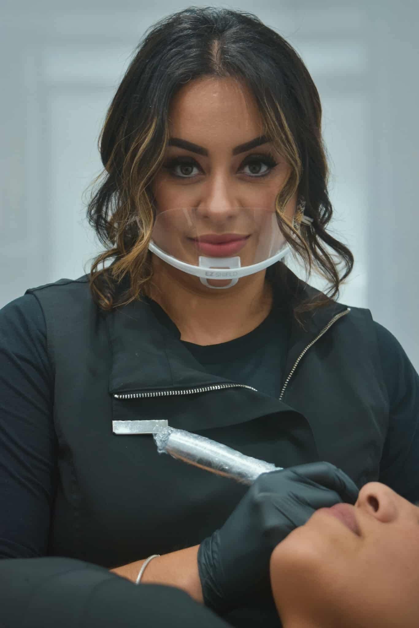 Woman in black gloves and face shield performing a lip procedure in a bright room.