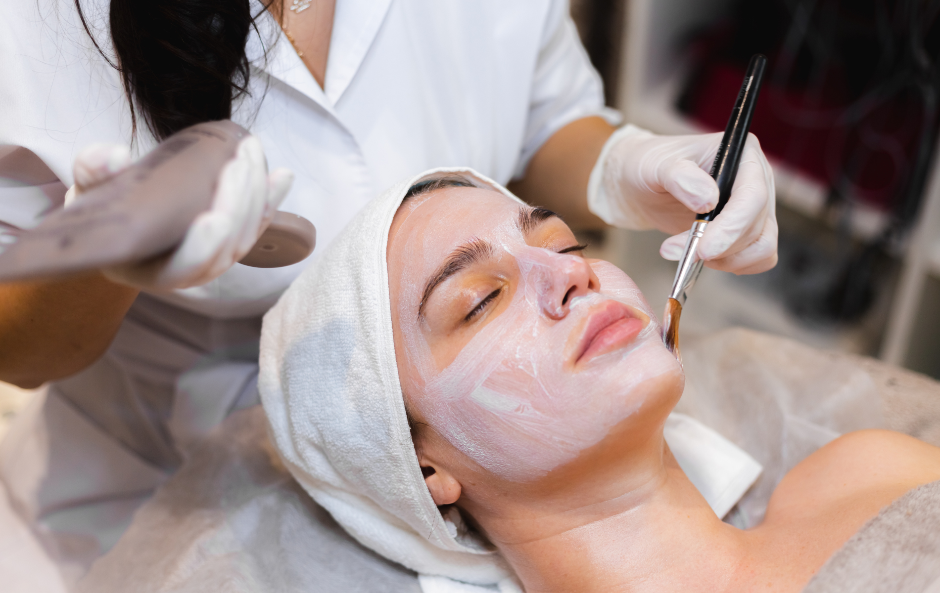 Facial treatment: Woman with face mask, esthetician applying product with brush in spa setting.