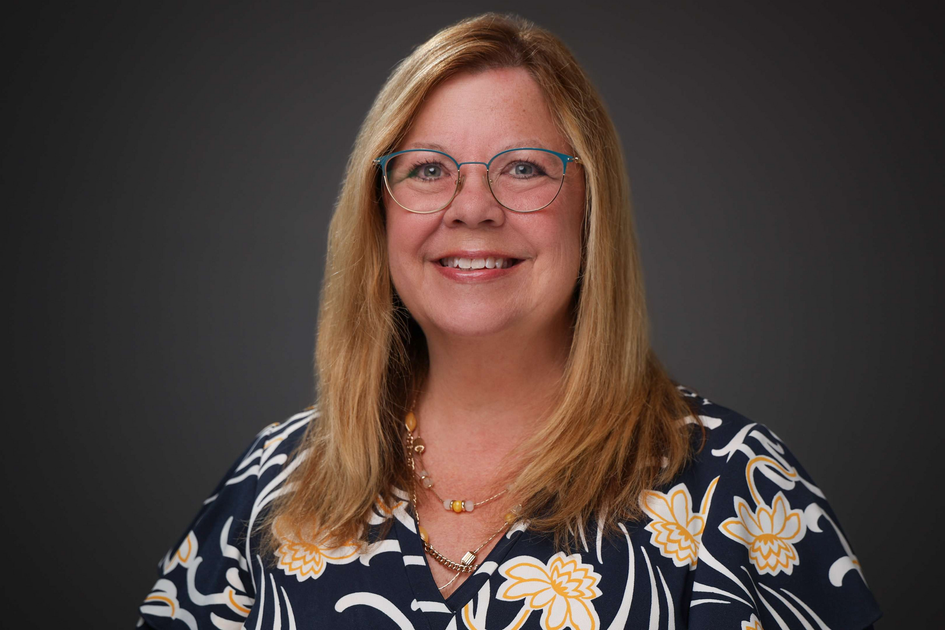 Smiling woman in glasses wearing a navy floral blouse against a dark gray background