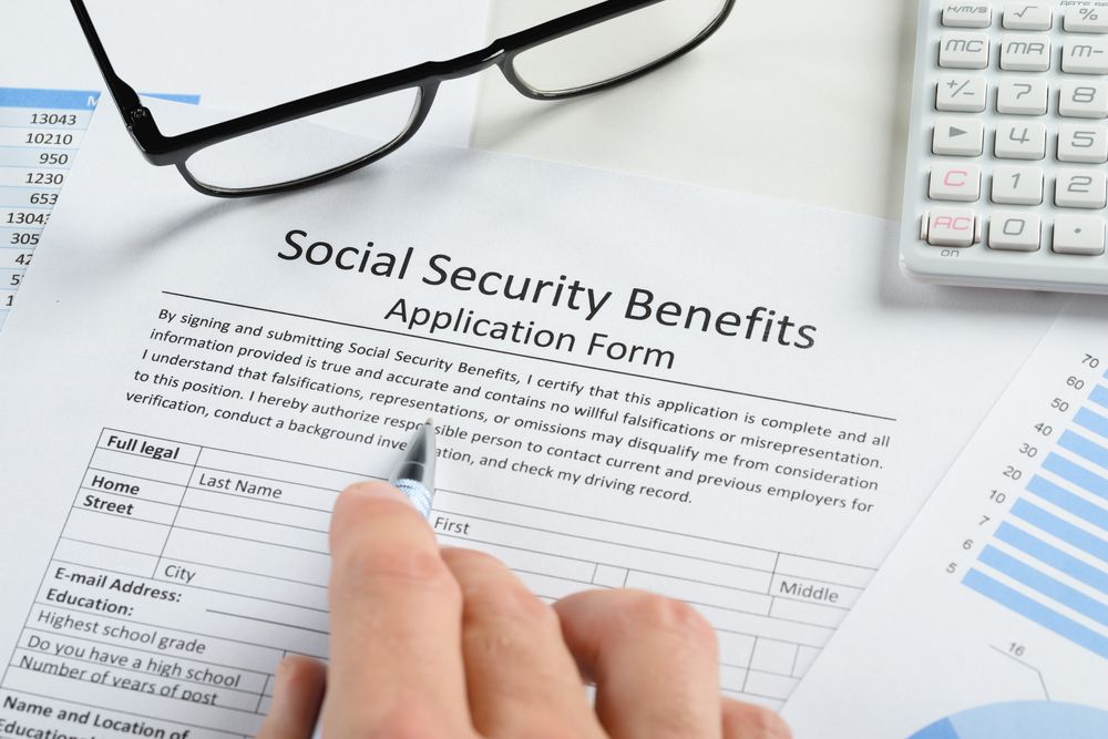 Social Security Benefits application form with hand, eyeglasses, calculator, and paperwork on desk