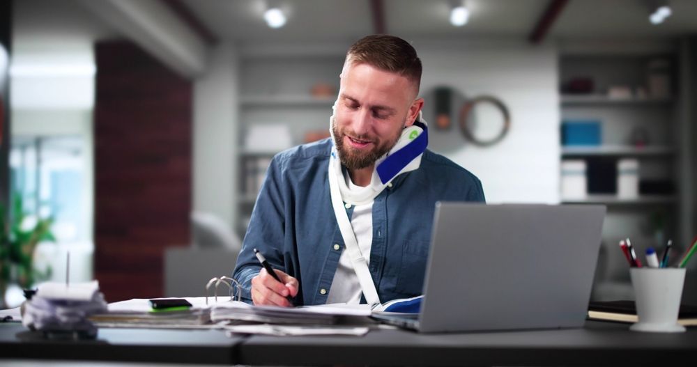 Man smiling at a desk, writing on paper beside a laptop in a modern office.