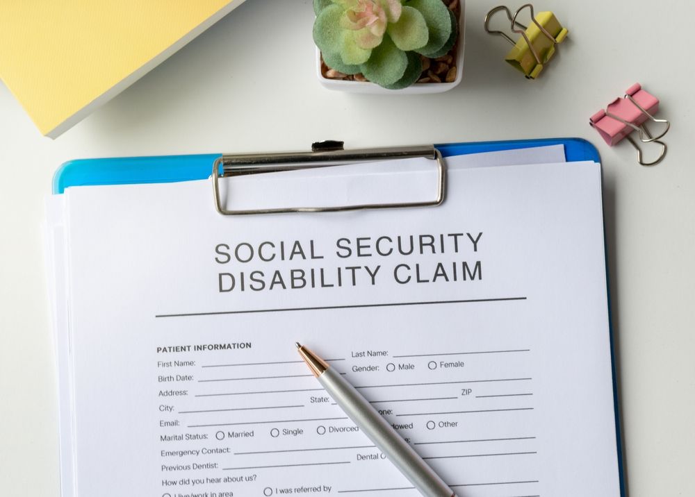 Clipboard with “Social Security Disability Claim” form and pen on a desk with a plant and clips.