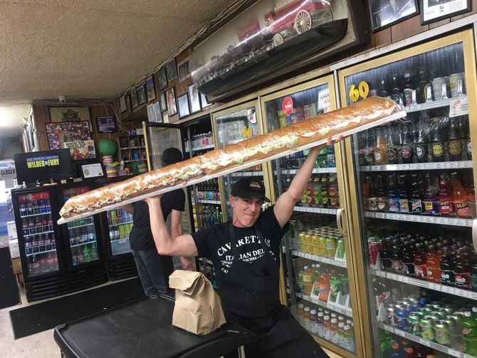 Person in a convenience store holding a giant baguette overhead, with shelves of drinks behind them.