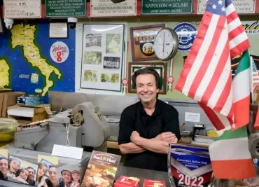 Smiling man behind a counter with souvenirs, a clock, posters, and a large American flag in a shop.