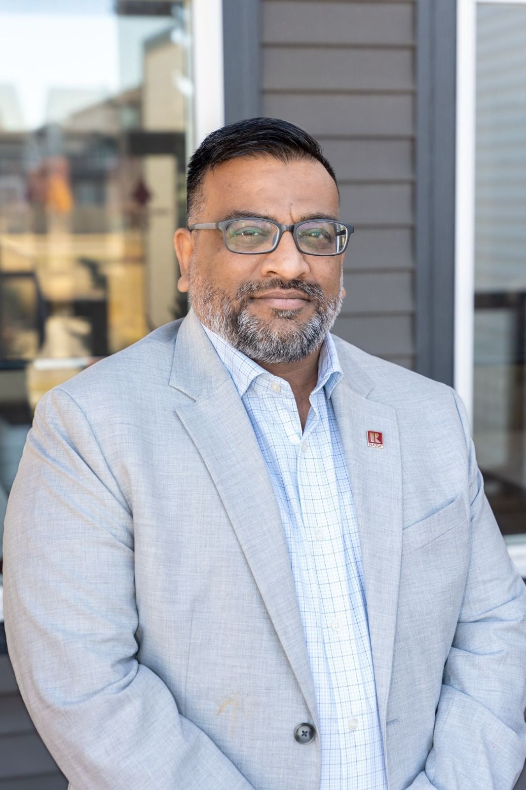 Man in a light gray blazer, collared shirt, and glasses, standing outdoors, smiling.