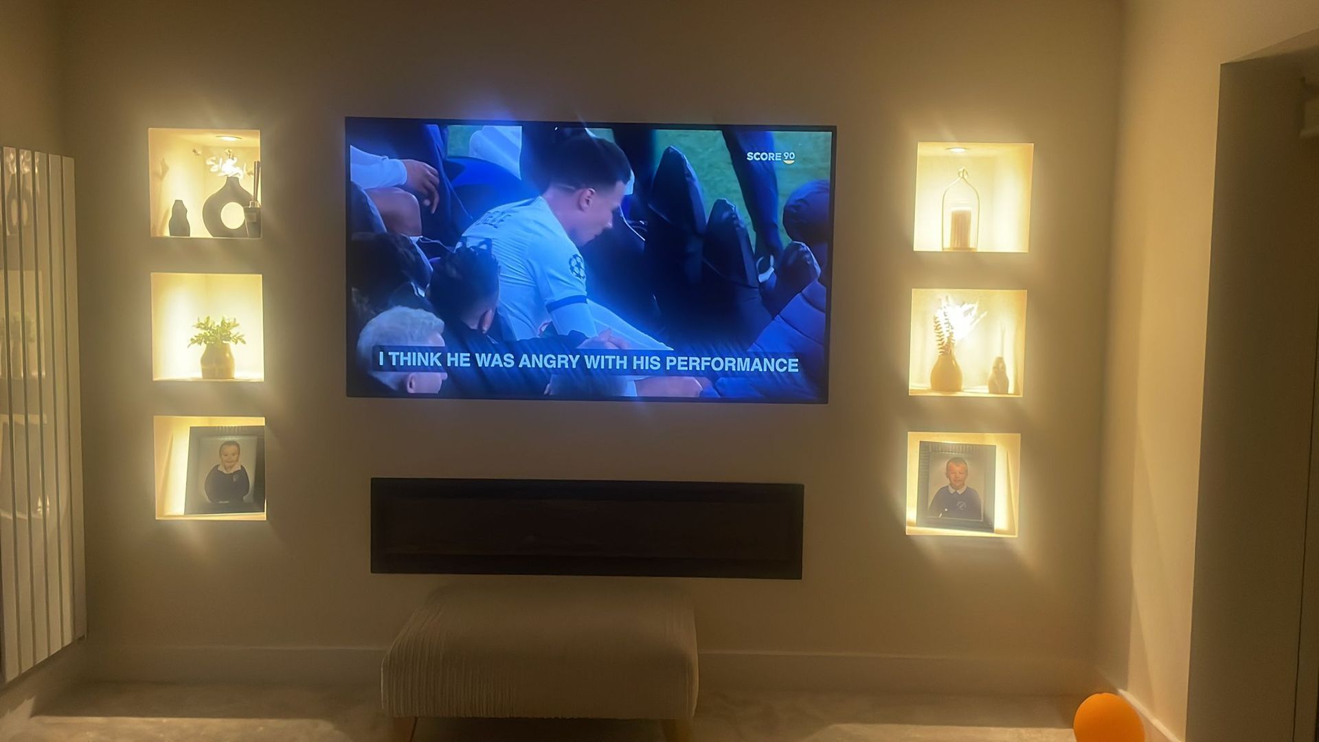 a picture of a media wall after it has been built and painted white with the TV and fireplace installed and turned on. There are also downlighters in the shelves.