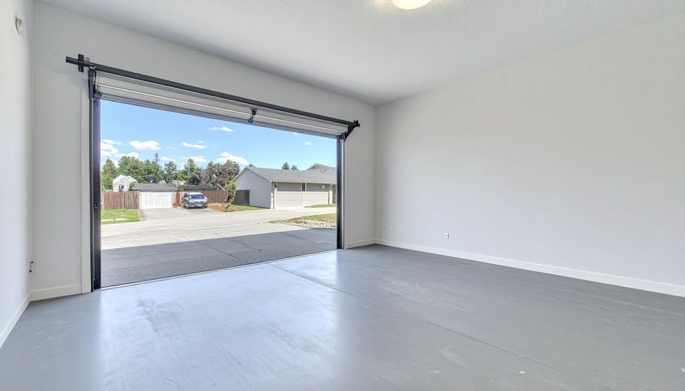 Empty garage interior with open garage door overlooking a street with houses and a blue sky. Gray floor and white walls.