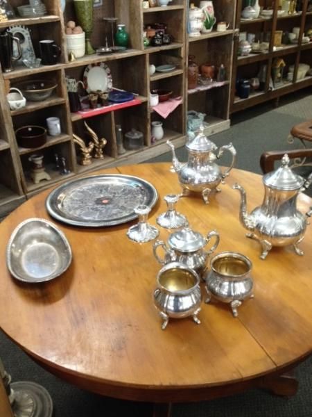 Silver tea set on a wooden table in a shop, with trays and shelves of assorted items in the background