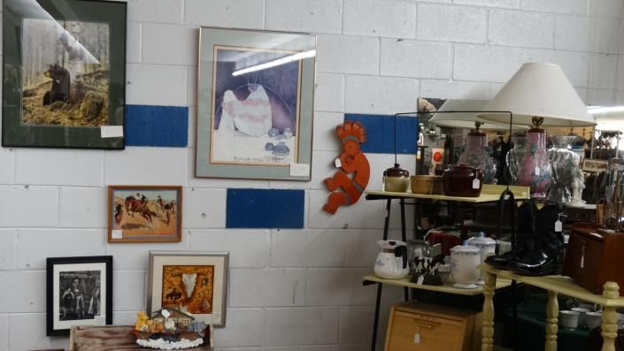 Wall of framed art above a shelf with lamps and glassware in a white room