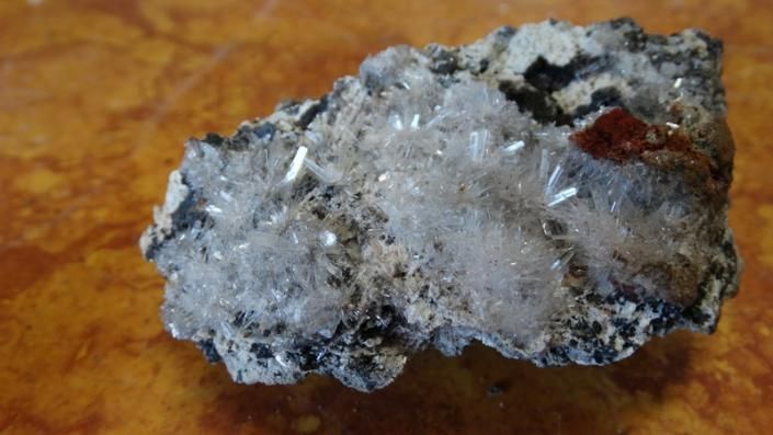 White crystalline mineral cluster on a brown surface, with dark rock and a small reddish patch.