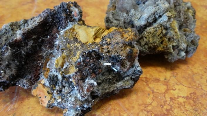 Three rough dark rocks with yellow and gray mineral deposits on a warm-toned surface