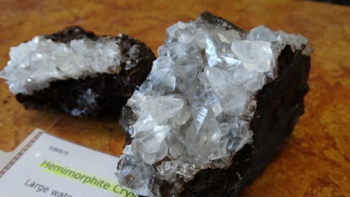 Two clear crystal clusters on dark rock, one larger specimen in the foreground with a small label card.