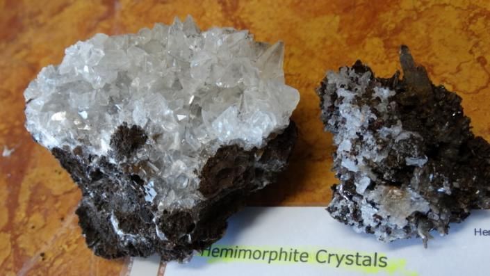 Two rough hemimorphite crystal rocks, one white and one dark gray, on a wooden surface