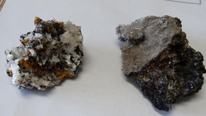 Two rough mineral rocks on a white surface, gray, black, and white with specks of orange.