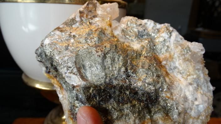 Close-up of a rough gray-and-gold rock held in a hand, with a white bowl in the background