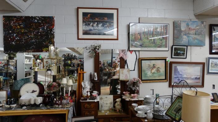 Cluttered room wall with many framed paintings, mirrors, and assorted decorative objects on shelves and tables