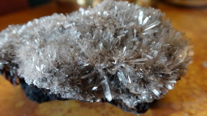 Silver-gray crystal cluster on dark rock against a warm wooden background