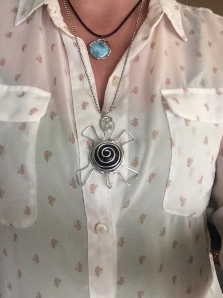 Person wearing a white patterned shirt with layered necklaces and a large spiral pendant.