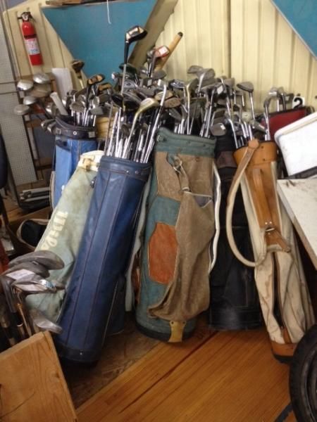 Golf clubs and bags crowded together in a storage corner on a wooden floor