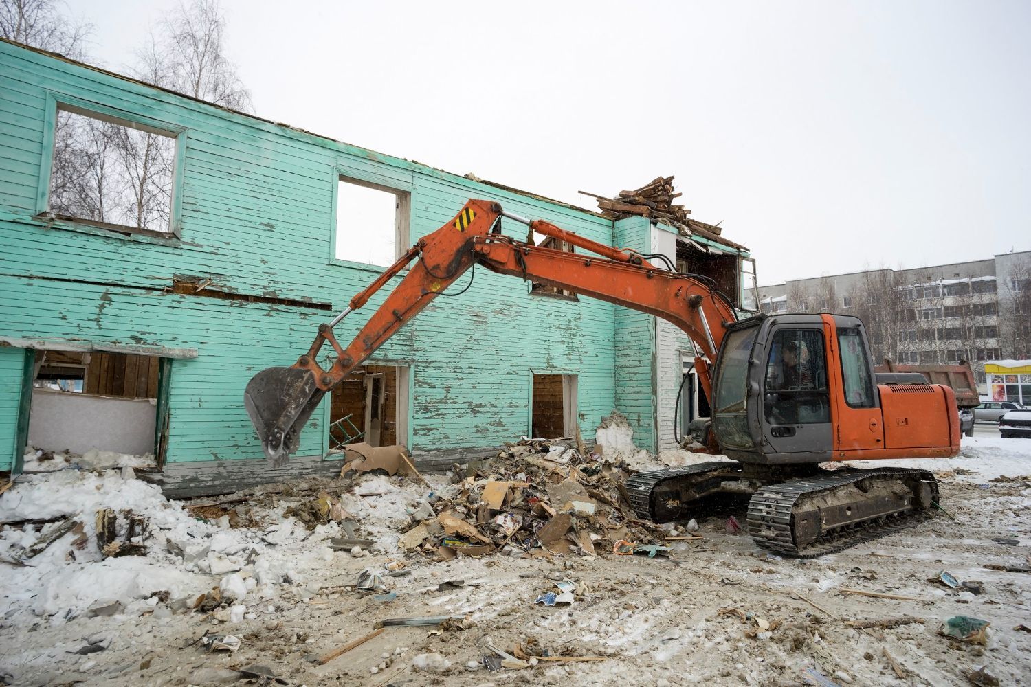 An orange excavator demolishes a teal-painted building in a snowy environment. Debris litters the ground.