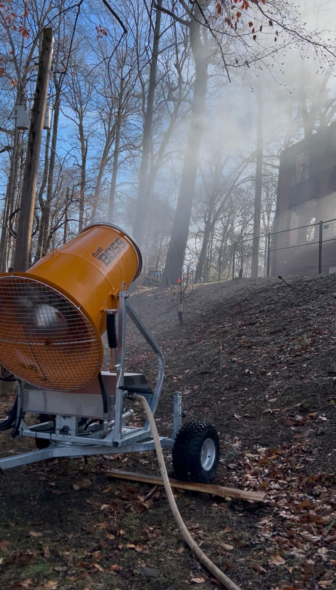 A snowmaking machine sprays mist into the air on a hillside with leafless trees.