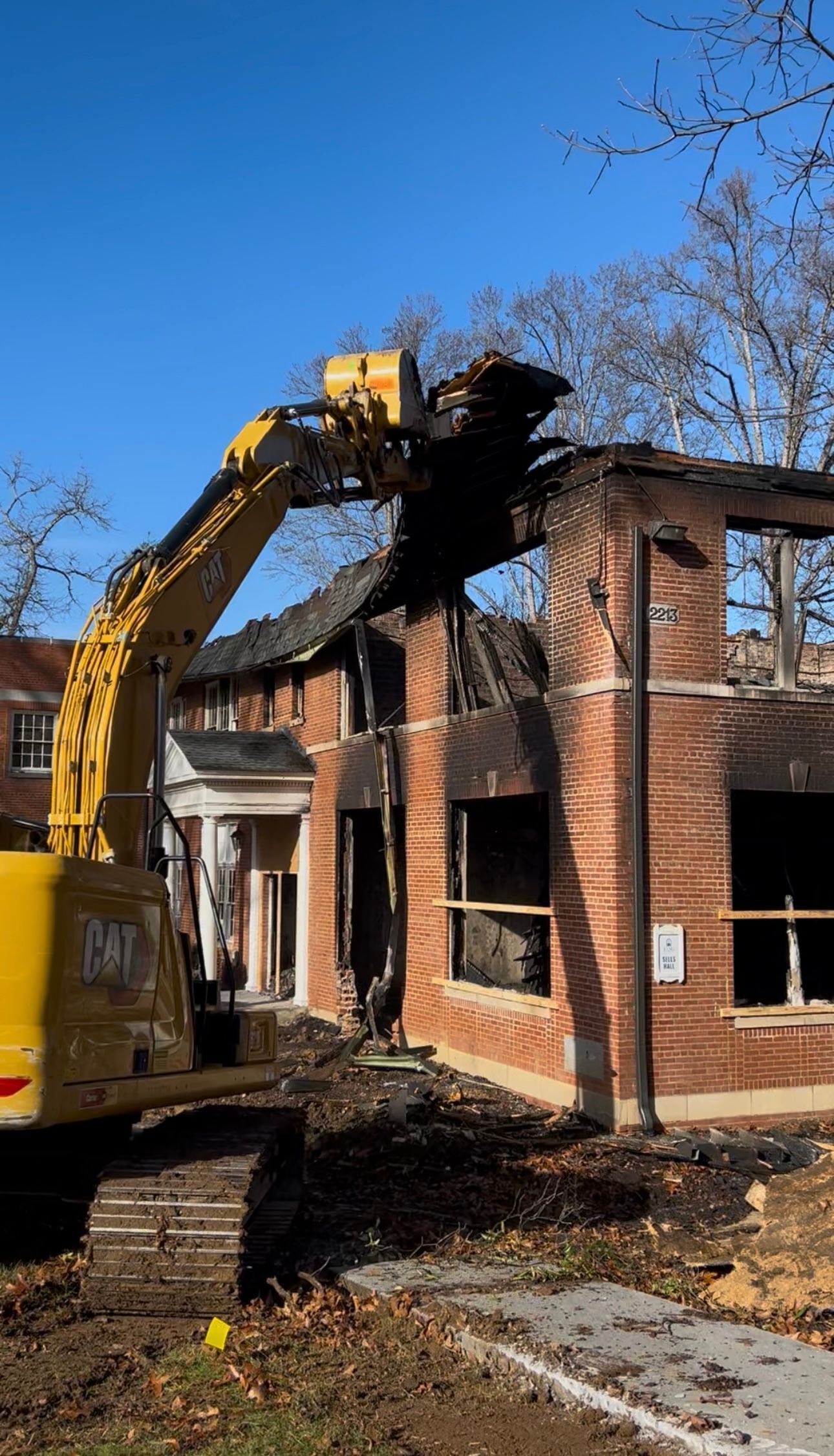 Excavator demolishing a two-story brick building with fire damage to the roof and windows.