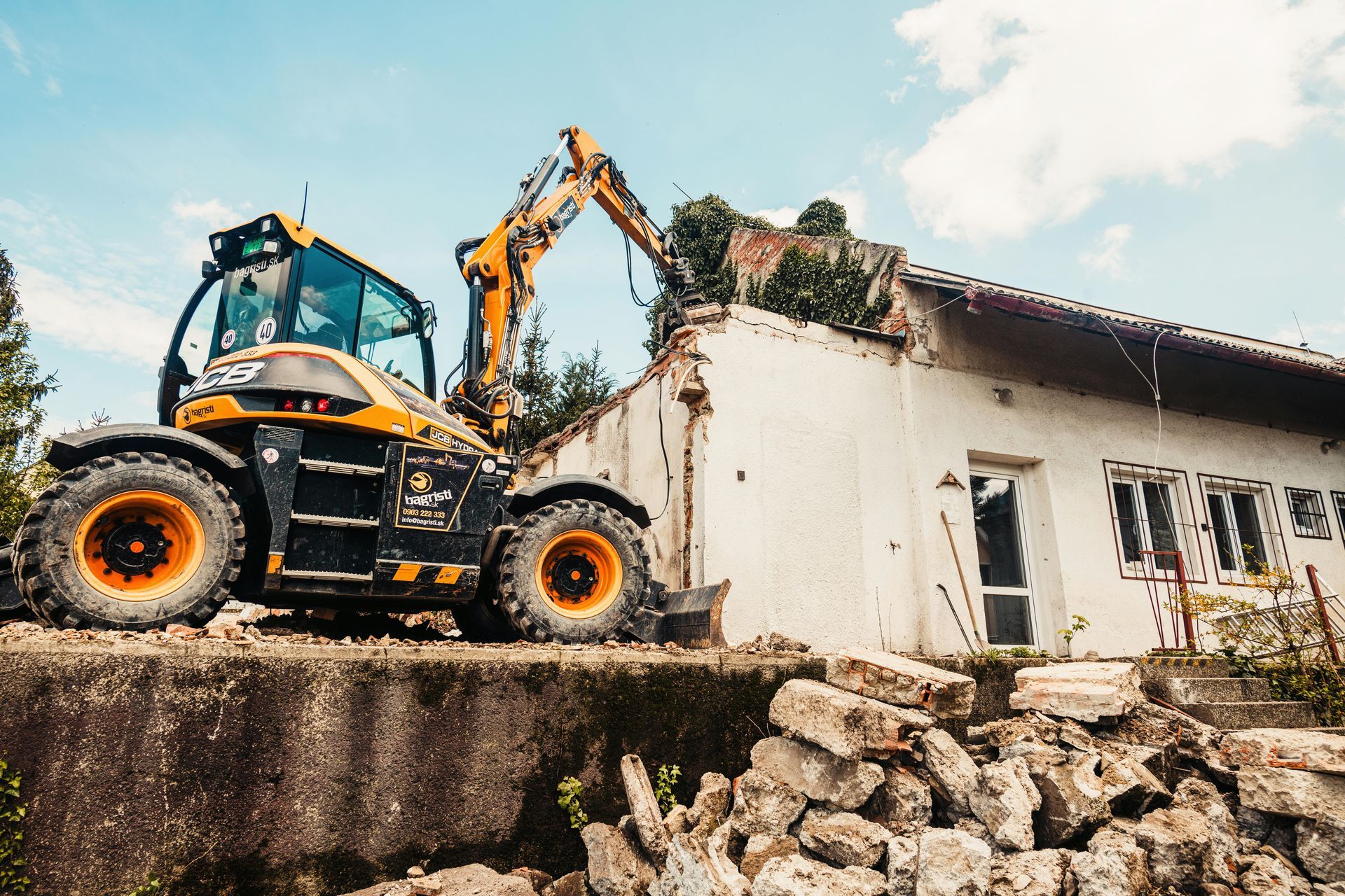 Excavator demolishing a white house beside a rubble pile