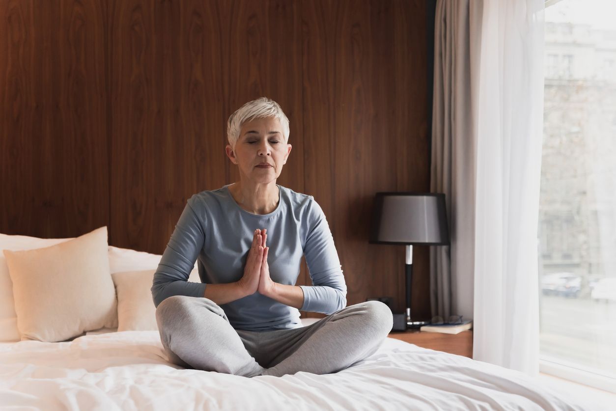 Woman meditating on a bed with hands in prayer position. Brown headboard, lamp, and window visible.