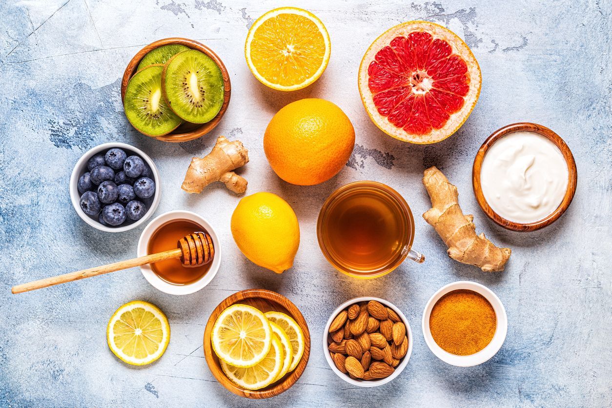 Assortment of immune-boosting foods: fruits, berries, nuts, ginger, honey, tea, and yogurt on a blue surface.