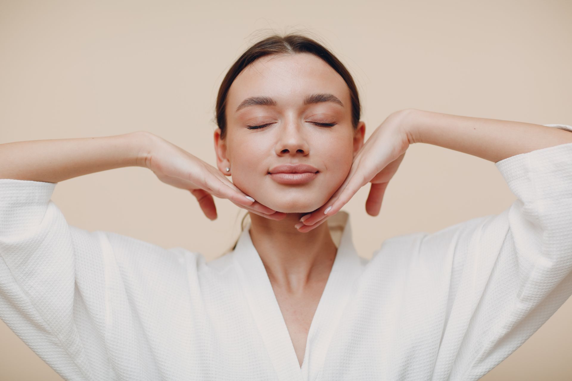 Woman in white robe, eyes closed, hands framing her face, beige background.