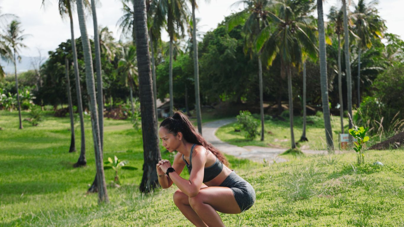 Woman doing squats outdoors, surrounded by palm trees and greenery.