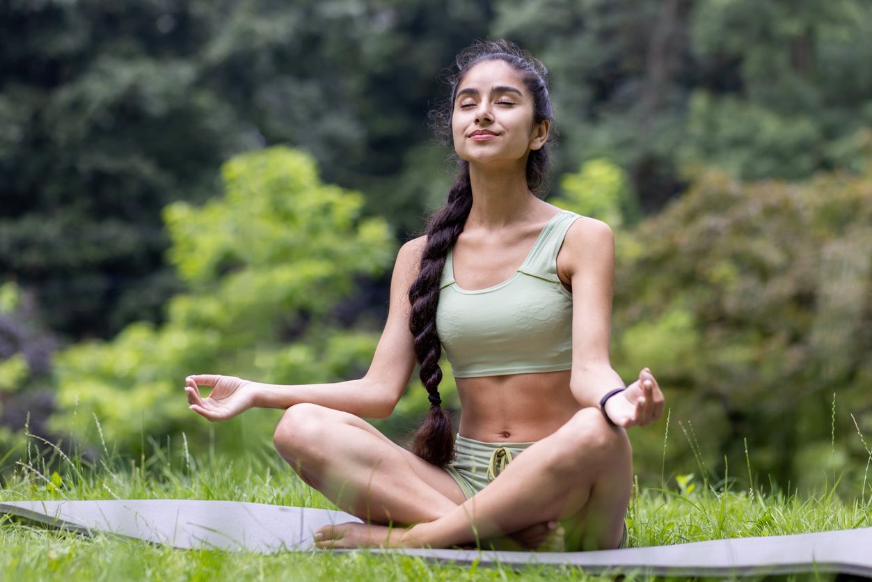 Woman meditating outdoors in a park, eyes closed, hands in prayer pose, wearing green activewear.
