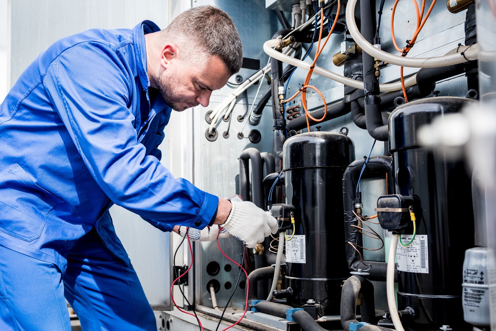 Photograph of a technician in blue coveralls working on industrial refrigeration equipment with multiple black compressors, pipes, and wiring. Photograph of a technician in blue coveralls working on industrial refrigeration equipment with multiple black compressors, pipes, and wiring.